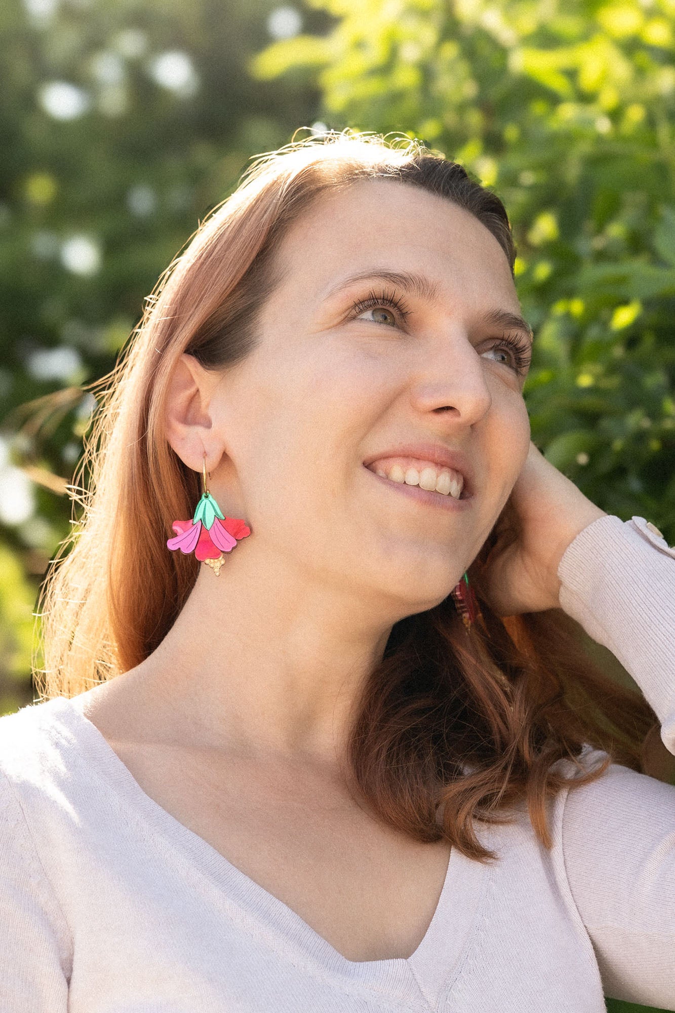 A woman with light brown hair smiles and looks upward, standing outdoors in sunlight. She wears a light long-sleeve top and handcrafted Hibiscus Earrings, with greenery and trees in the background.
