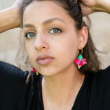 A young woman with light eyes and wavy brown hair poses with her hands behind her head, wearing a black top and handcrafted, colorful Hibiscus Earrings, looking directly at the camera against a neutral background.