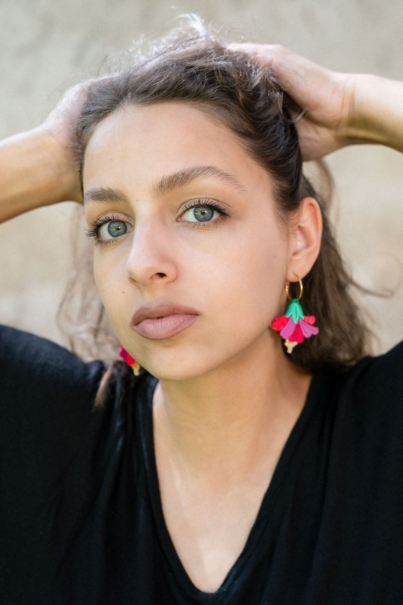 A young woman with light eyes and wavy brown hair poses with her hands behind her head, wearing a black top and handcrafted, colorful Hibiscus Earrings, looking directly at the camera against a neutral background.