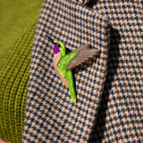 A close-up of a handcrafted hummingbird brooch, made from premium materials, pinned to a houndstooth-patterned coat with a green turtleneck sweater visible underneath.