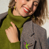 A smiling woman in a green turtleneck and plaid jacket stands against a light wall, wearing handcrafted Hummingbird Earrings and matching lapel jewelry. Sunlight highlights her face as she closes her eyes, radiating joy and sustainable craftsmanship.