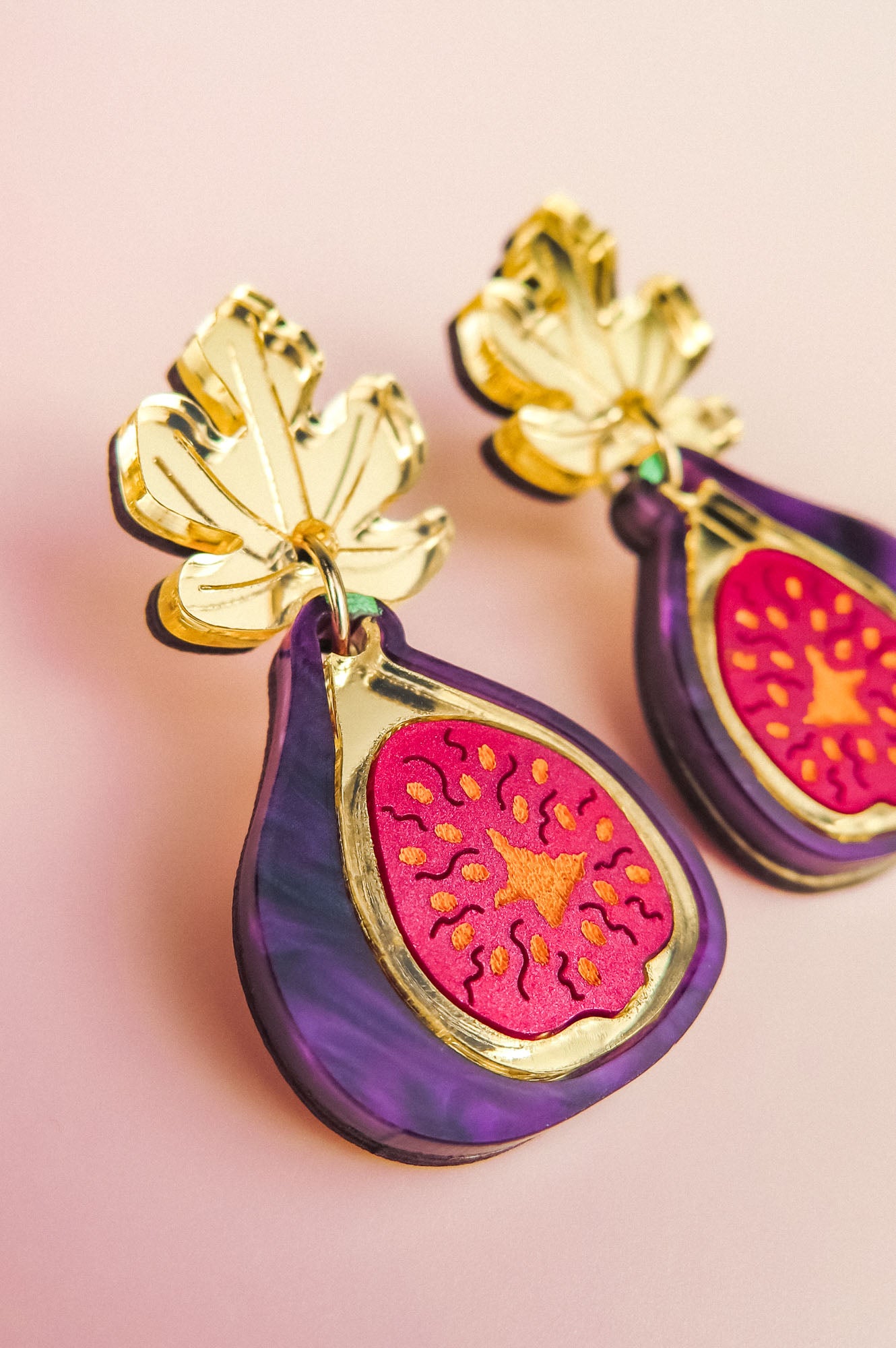 A close-up of handcrafted Fig Earrings shaped like figs, featuring gold mirror leaves at the top, vibrant purple edges, and a bright pink and orange fig center, set against a soft pink background.