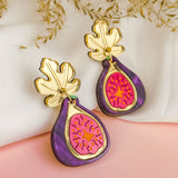 A pair of handcrafted Fig Earrings with gold mirror leaves, purple edges, and a pink center, displayed on a soft white and pink background with dried flowers.