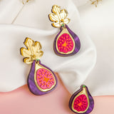 Three handcrafted Fig Earrings, featuring gold mirror leaves and vibrant purple and pink detailing, are displayed on white fabric and a pink surface, with dried foliage in the background.