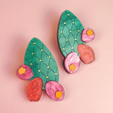 A pair of statement earrings shaped like green opuncia cactus pads with pink and orange flowers, decorated with small yellow dots, displayed on a light pink background.