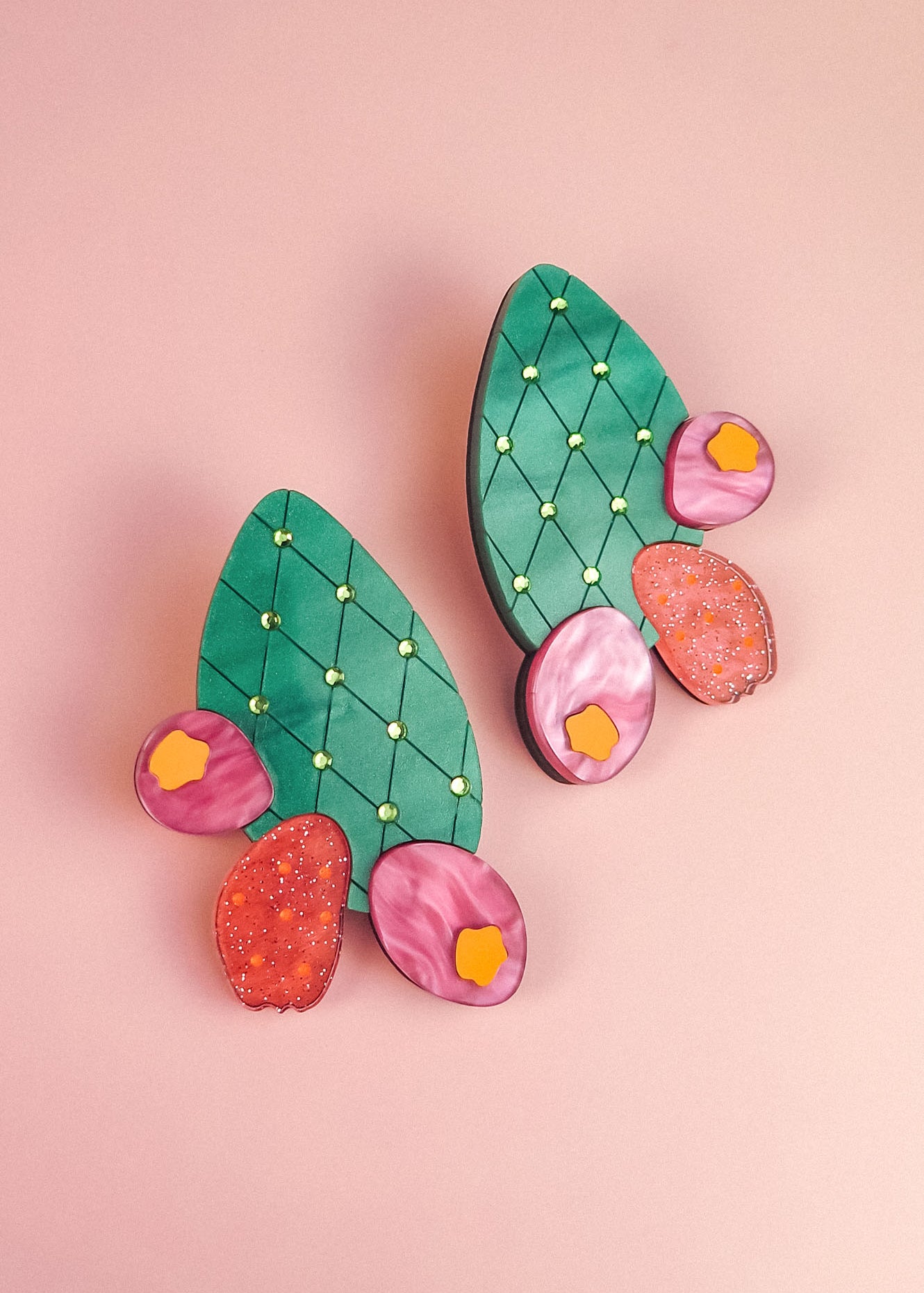 A pair of statement earrings shaped like green opuncia cactus pads with pink and orange flowers, decorated with small yellow dots, displayed on a light pink background.