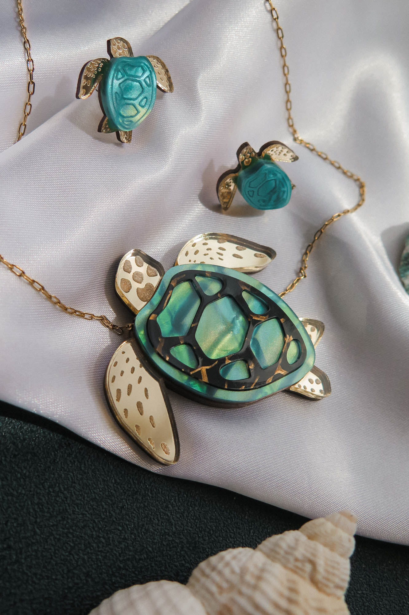 A handcrafted gold jewelry set shaped like sea turtles with green and cream details is displayed on white satin fabric. The set includes a Sea Turtle Necklace, a ring, and earrings. A shell is placed nearby.