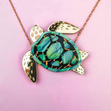 This handcrafted jewelry piece is a gold chain Sea Turtle Necklace, featuring a turtle pendant with a green and black patterned shell and elegant gold accents, beautifully set against a pink background.