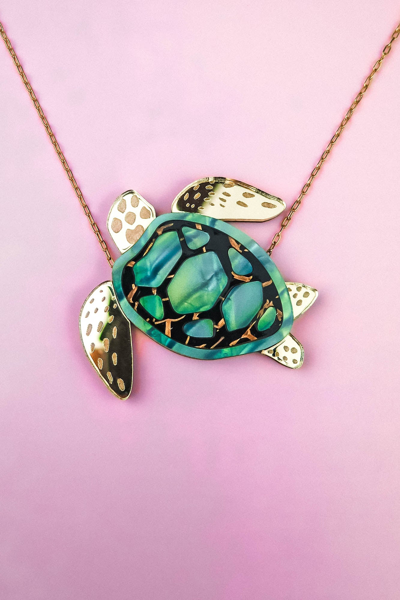 This handcrafted jewelry piece is a gold chain Sea Turtle Necklace, featuring a turtle pendant with a green and black patterned shell and elegant gold accents, beautifully set against a pink background.