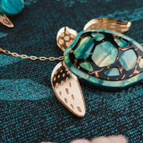 A close-up of a gold and turquoise Sea Turtle Necklace lies on a textured blue surface, with seashells and other handcrafted jewelry featuring turtle-themed designs nearby.