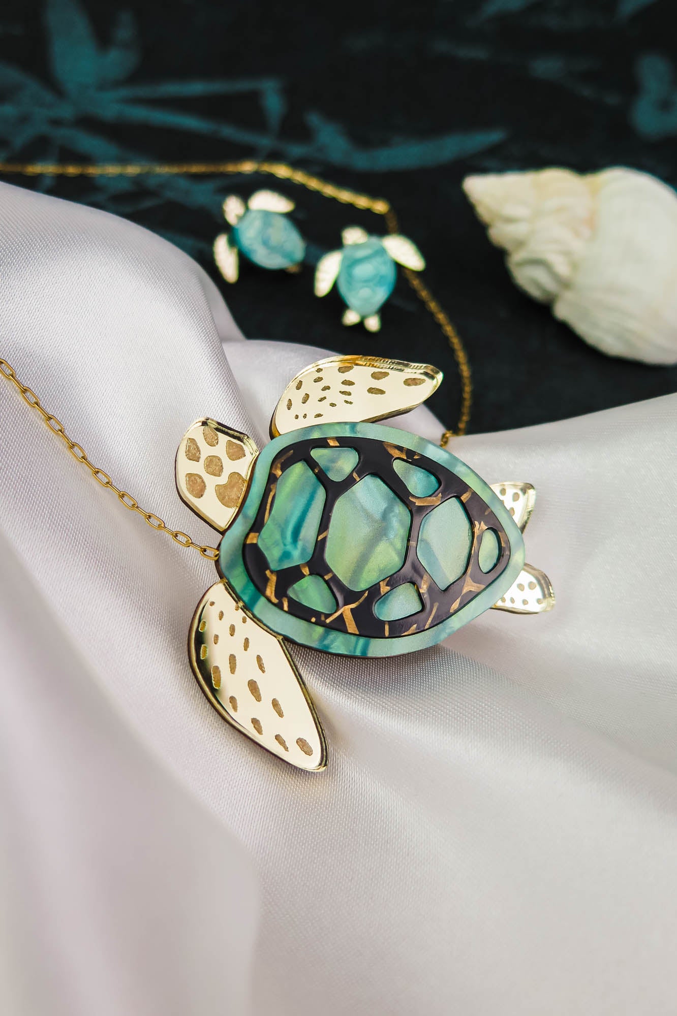A gold and green Sea Turtle Necklace pendant, handcrafted jewelry with intricate details, is displayed on silky white fabric alongside a matching smaller turtle necklace in the background and a seashell nearby.