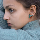 A young person wearing a teal knit sweater looks to the side, showing a Kingfisher Stud Earring. Their hair is pulled back, and their expression appears calm and thoughtful, embracing an eco-friendly, sustainably made accessory.