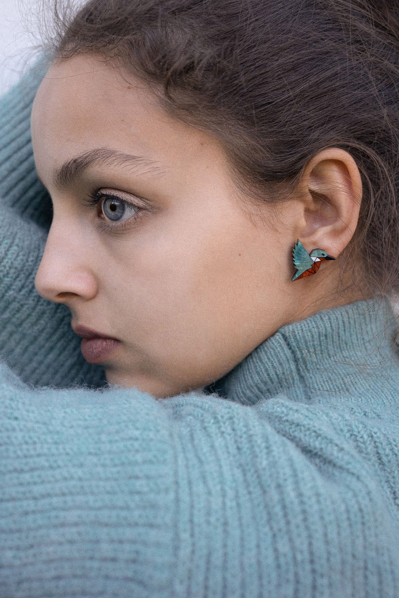 A young person wearing a teal knit sweater looks to the side, showing a Kingfisher Stud Earring. Their hair is pulled back, and their expression appears calm and thoughtful, embracing an eco-friendly, sustainably made accessory.
