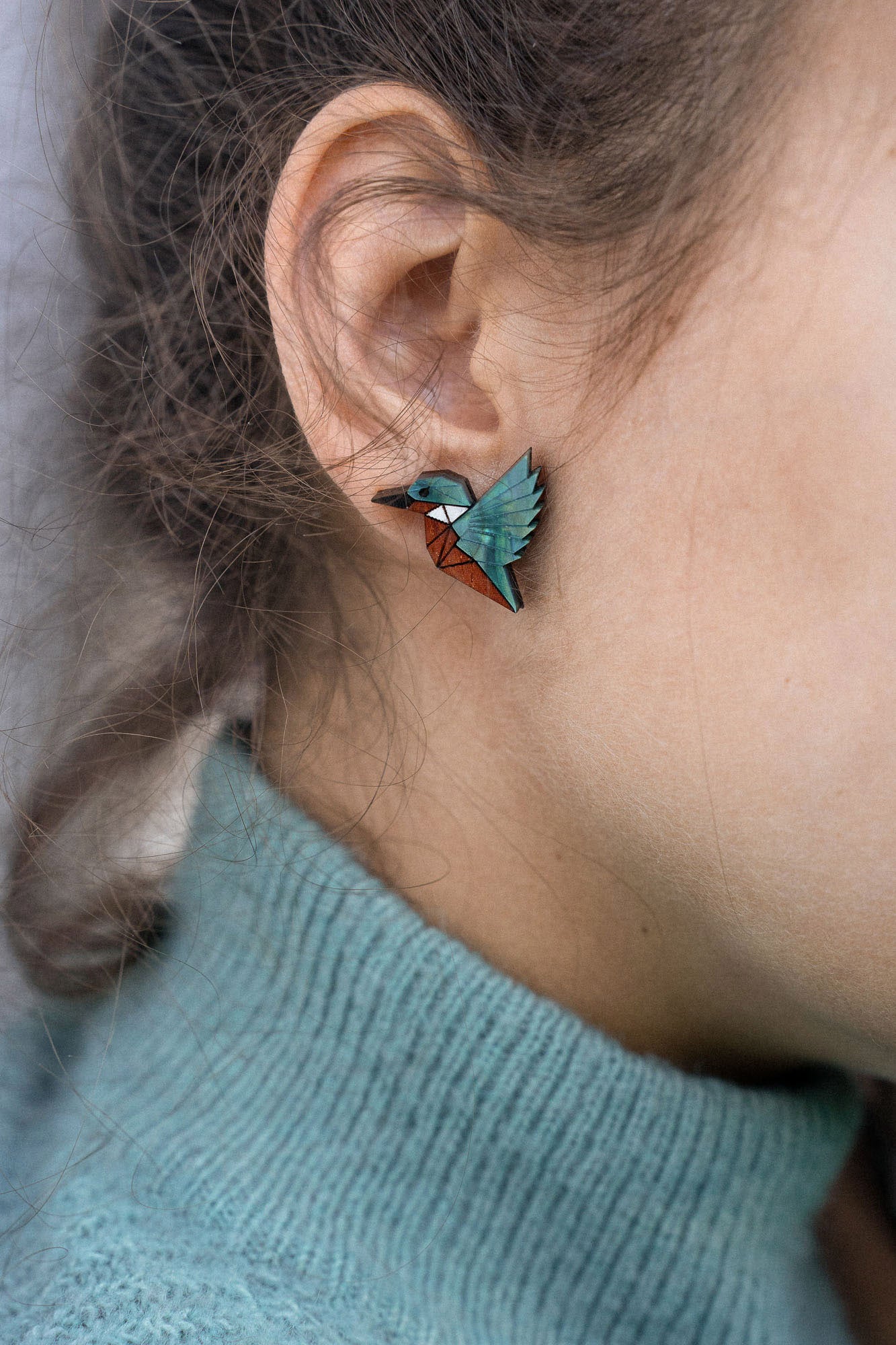Close-up of a person wearing a turquoise sweater and eco-friendly Kingfisher Stud Earrings, shaped like a hummingbird, with the focus on their ear and earring. The background is softly blurred.