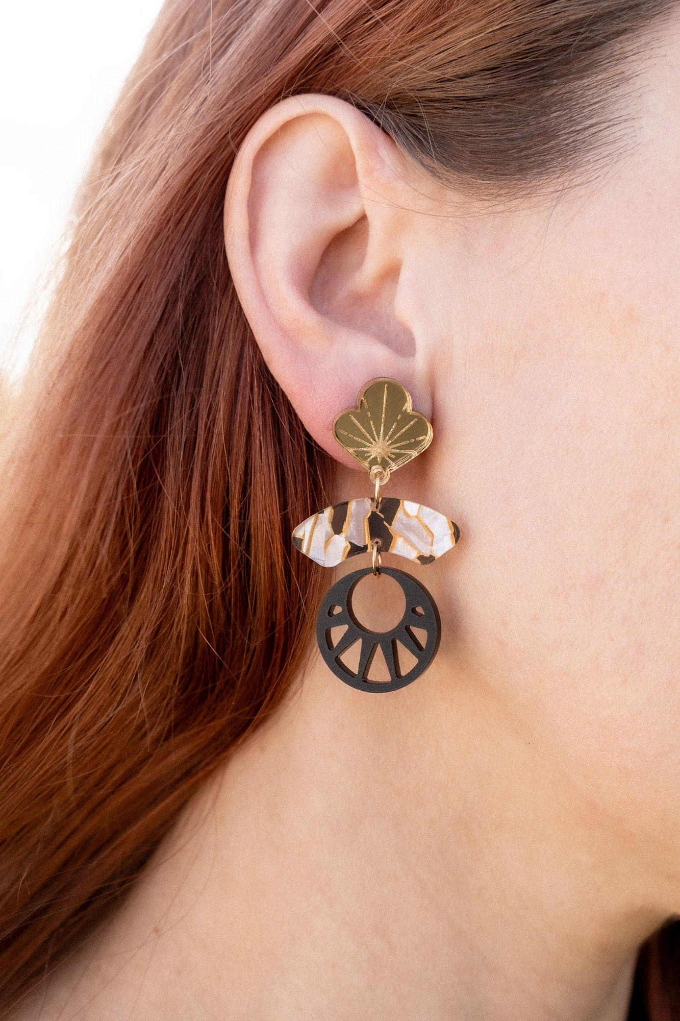 Close-up of a person with long auburn hair wearing handcrafted Kintsugi Earrings featuring a gold leaf, a clear crystal, and a black circular charm with a cut-out sunburst design inspired by Japanese art.