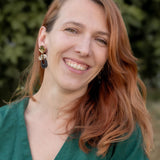 A woman with long, reddish-brown hair smiles warmly at the camera. She is wearing a green top and striking handcrafted Kintsugi Earrings. The blurred greenery in the background suggests an outdoor setting.