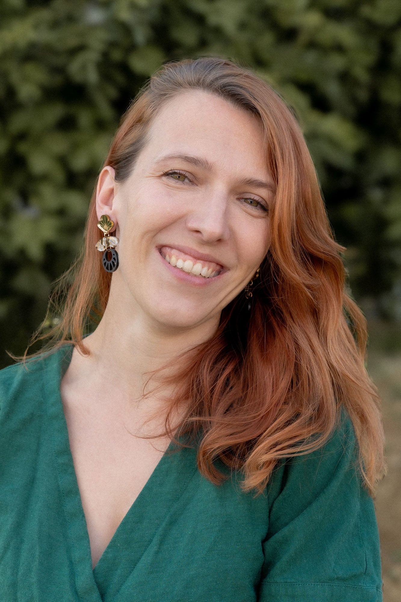A woman with long, reddish-brown hair smiles warmly at the camera. She is wearing a green top and striking handcrafted Kintsugi Earrings. The blurred greenery in the background suggests an outdoor setting.