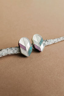 Handmade geometric stud earrings with silver, lavender, and teal sections rest on a textured, light gray branch against a beige background—a perfect touch of minimalist jewelry.