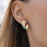 Close-up of a womans ear wearing a geometric, gold-toned stud earring with pastel accents. This piece of minimalist jewelry stands out against the softly blurred background, highlighting its unique handmade design and part of her hair and face.