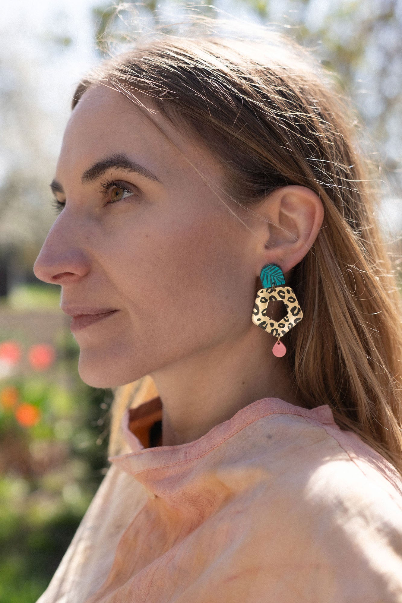 A woman with light brown hair gazes to the left, wearing a peach top and striking Leopard Earrings crafted from premium materials. The background is outdoors with blurred greenery and flowers.