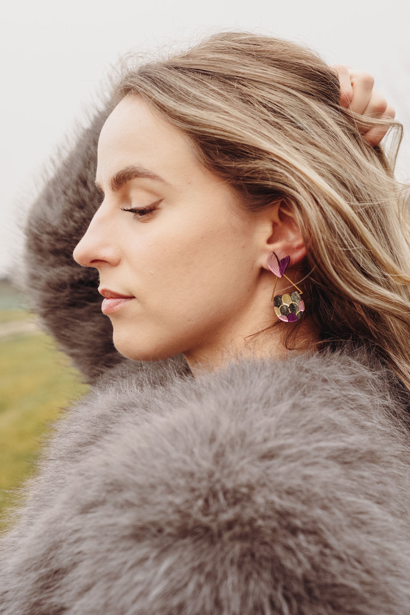 A woman with wavy blonde hair, wearing a fluffy gray coat and playful Mermaid Tail Earrings, stands outdoors with her eyes closed and one hand in her hair. The background is grassy with blurred trees—perfect for mermaid lovers.