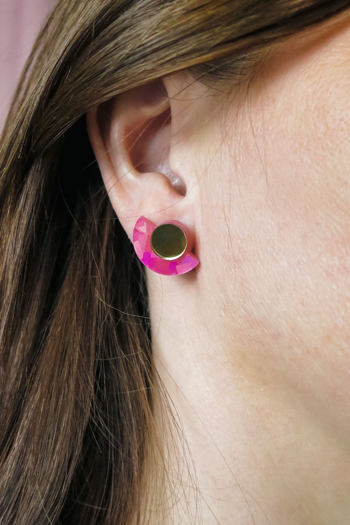 Close-up of a persons ear wearing lightweight Minimalist Geometric Stud Earrings, featuring a round gold stud and a pink, crescent-shaped accent. The person has straight brown hair tucked behind the ear.