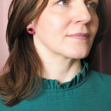 A woman with shoulder-length brown hair wearing a teal top with a ruffled collar and Minimalist Geometric Stud Earrings stands in front of pink curtains, looking slightly to the right with a neutral expression.