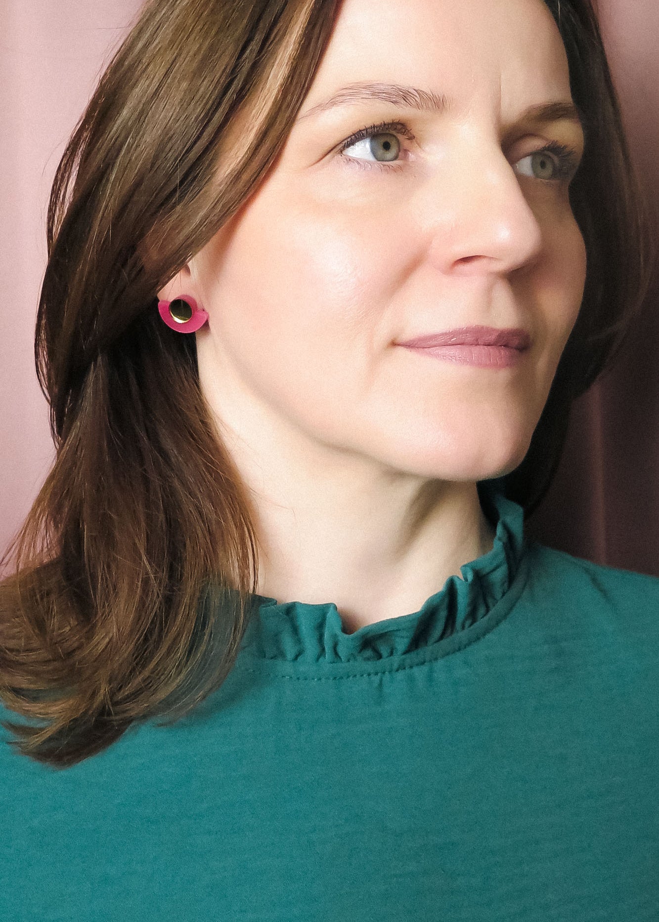 A woman with shoulder-length brown hair wearing a teal top with a ruffled collar and Minimalist Geometric Stud Earrings stands in front of pink curtains, looking slightly to the right with a neutral expression.
