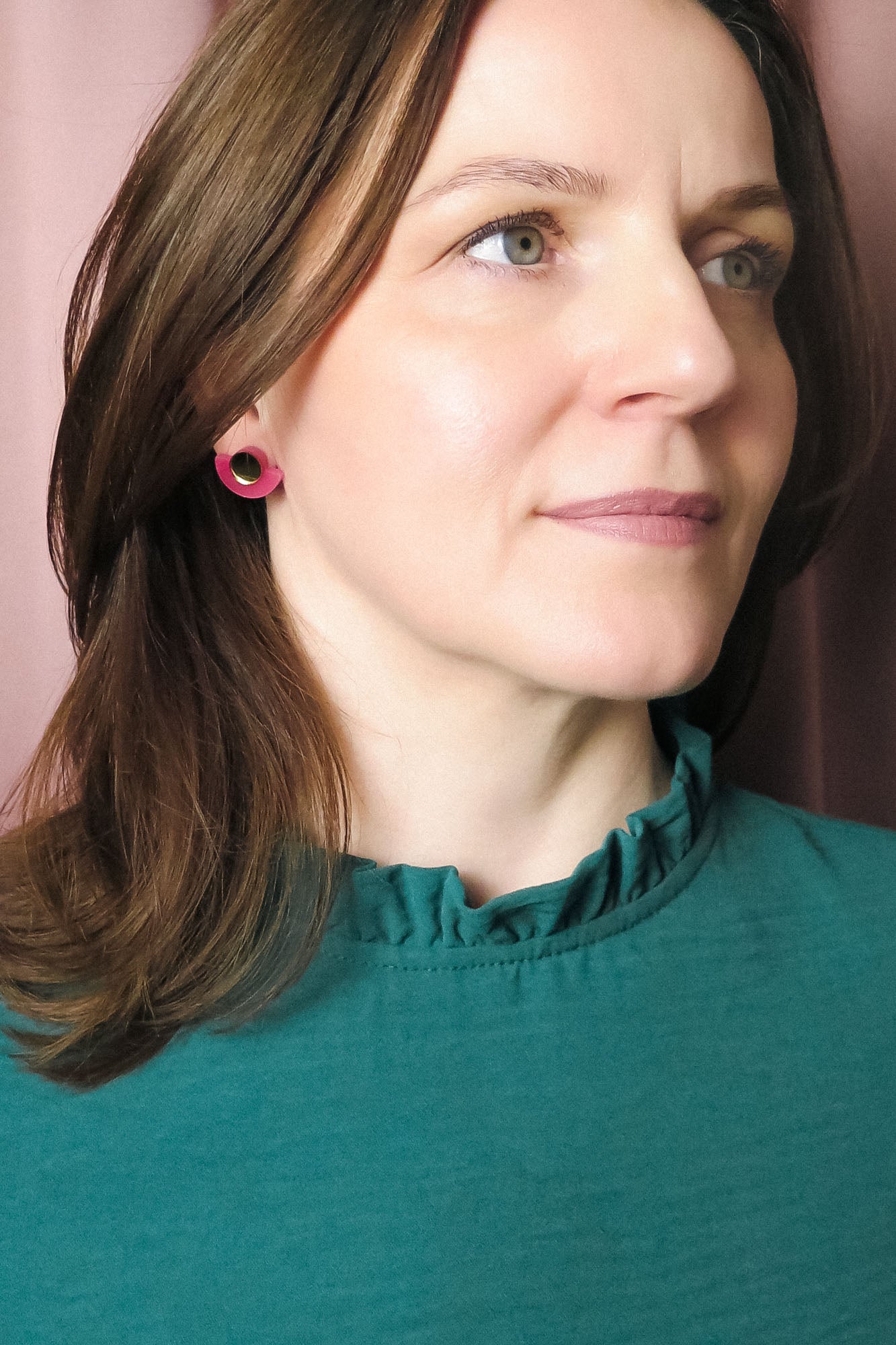 A woman with shoulder-length brown hair wearing a teal top with a ruffled collar and Minimalist Geometric Stud Earrings stands in front of pink curtains, looking slightly to the right with a neutral expression.