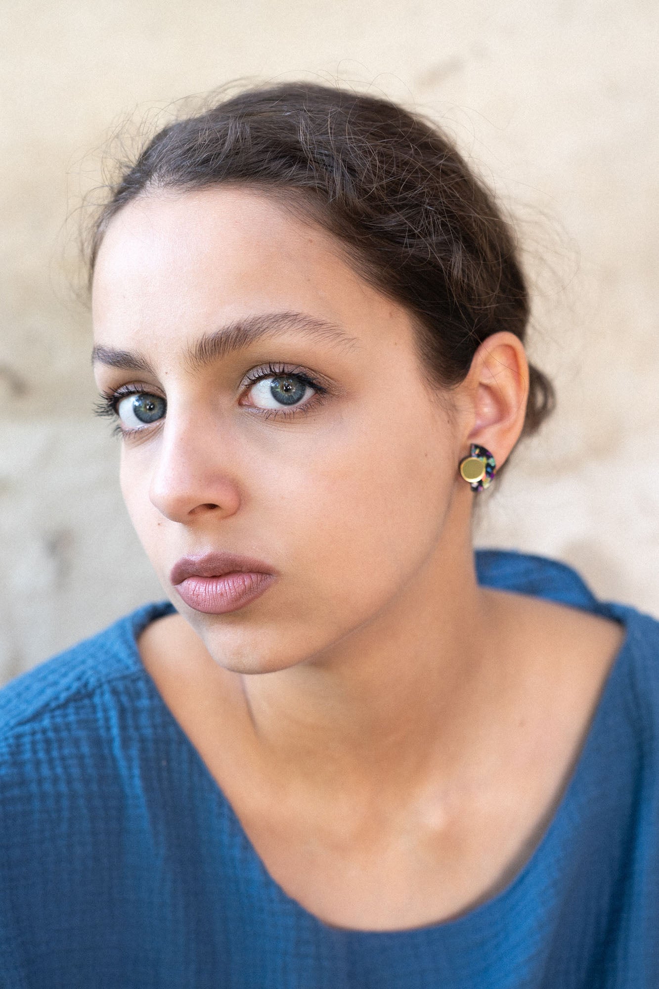 A young woman with light eyes and dark hair pulled back, wearing a textured blue top and hypoallergenic Minimalist Geometric Stud Earrings, gazes seriously at the camera against a neutral, slightly textured background.