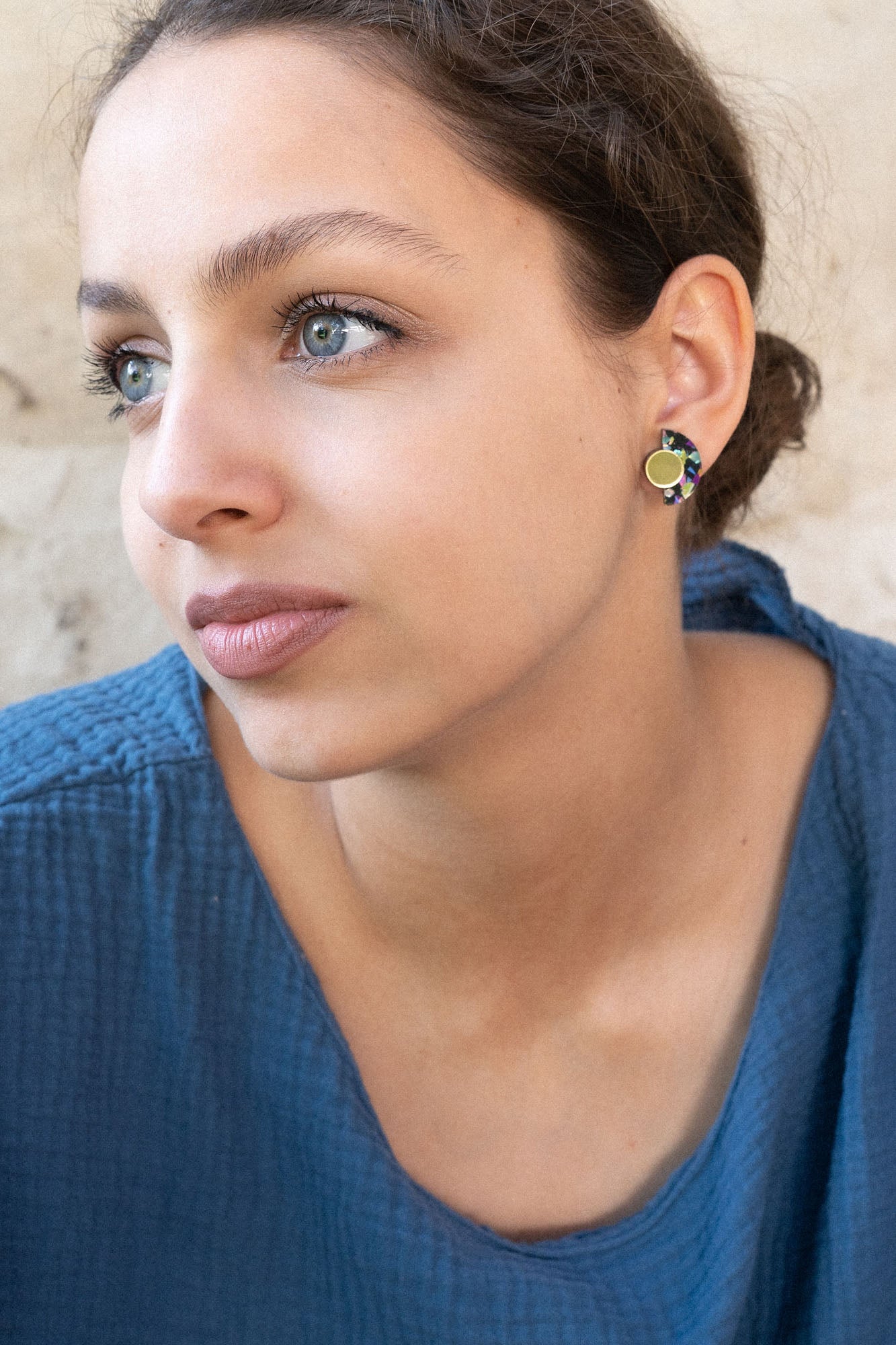 A young woman with light eyes and brown hair pulled back, wearing a textured blue top and lightweight Minimalist Geometric Stud Earrings, looks thoughtfully to the side against a beige, textured wall.
