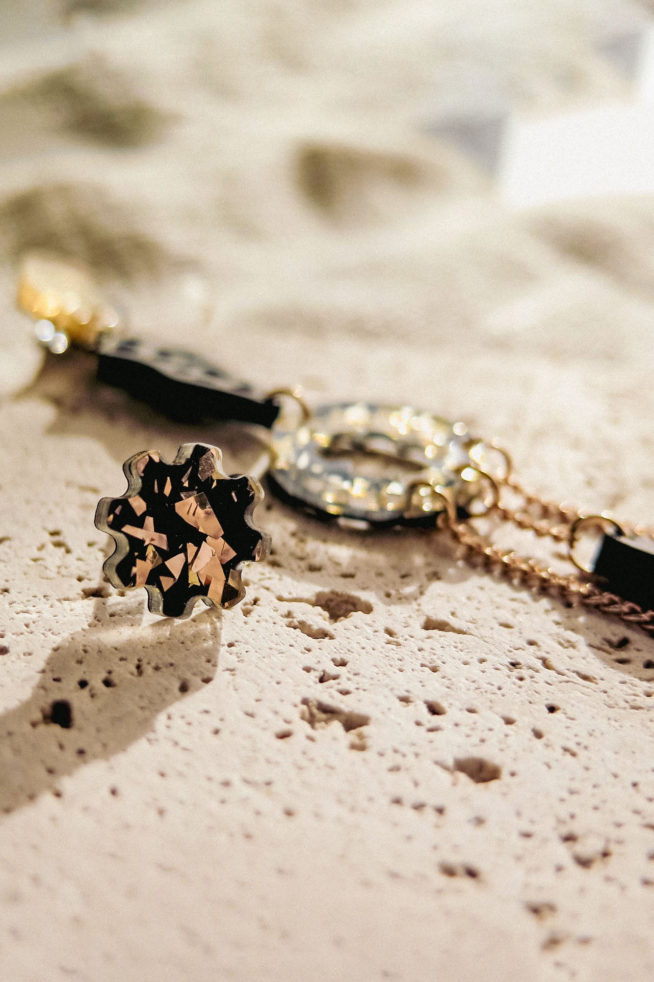 Close-up of a stylish gold bracelet and a matching floral-shaped ring with black and gold details, paired with lightweight handcrafted earrings, all arranged on a textured, light-colored stone surface in soft natural light.