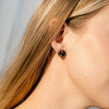 Close-up of a woman's ear with straight blonde hair, wearing a small, black and gold floral stud earring. These handcrafted earrings are lightweight, adding elegance without compromising comfort. The background is softly blurred in natural light.