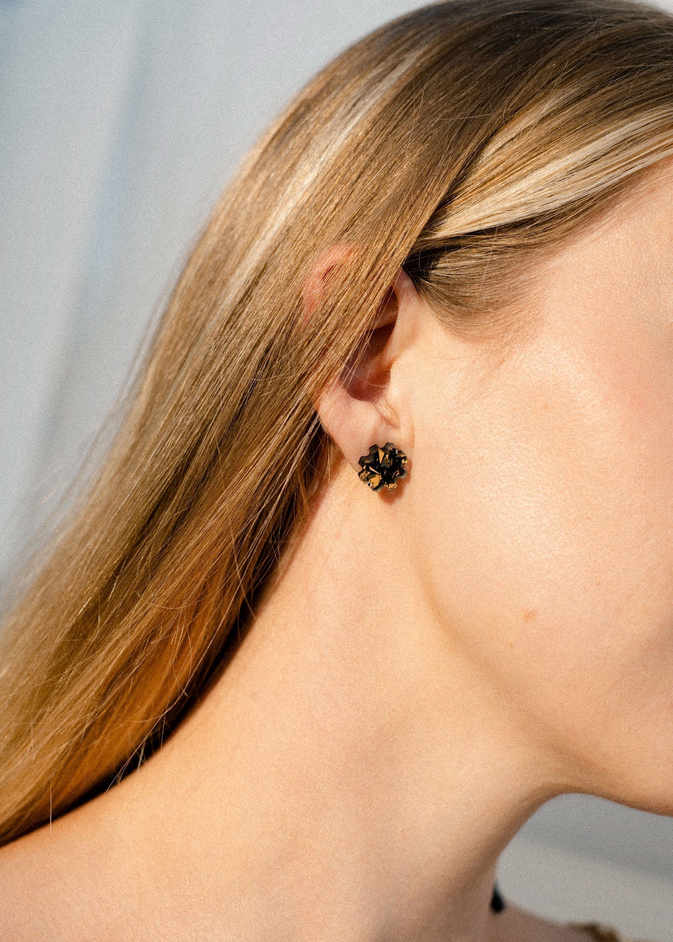 Close-up of a woman's ear with straight blonde hair, wearing a small, black and gold floral stud earring. These handcrafted earrings are lightweight, adding elegance without compromising comfort. The background is softly blurred in natural light.