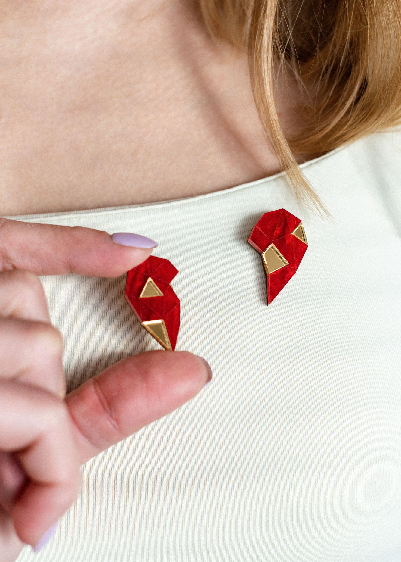 A person with long hair wears a cream top, holding a red and gold geometric One Love Halves Pin shaped like half a broken heart—the other handcrafted half is pinned to their shirt near the collarbone. Perfect as a romantic gift.