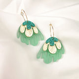 A pair of handcrafted dangling earrings with gold hooks, featuring layered designs in mint green, cream, and teal glitter, resembling abstract floral shapes for a touch of tropical elegance, displayed on a white fabric background.