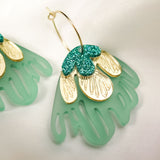 A close-up of a handcrafted gold hoop earring featuring green and gold petal-shaped charms with glittery accents, embodying tropical elegance, set on a soft white fabric background.