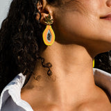 A woman with curly dark hair wears a white shirt and a colorful pair of Papaya Earrings, looking upward against a light background. These vibrant, handcrafted earrings add a playful touch to her modern style.