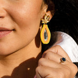 A woman wearing a white shirt shows off a gold ring with a dark blue stone and a colorful Papaya Earring, handcrafted in the shape of a papaya half.