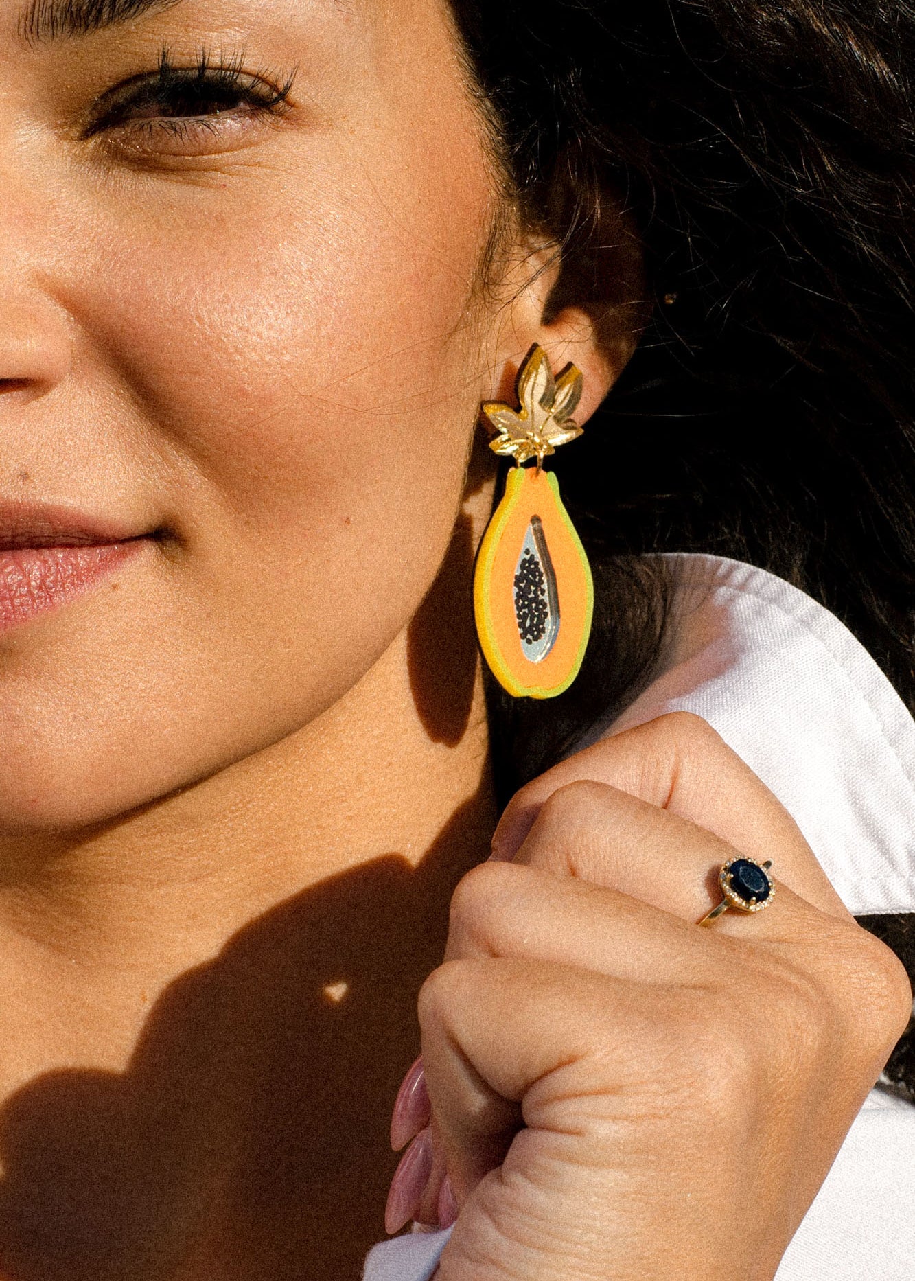 A woman wearing a white shirt shows off a gold ring with a dark blue stone and a colorful Papaya Earring, handcrafted in the shape of a papaya half.