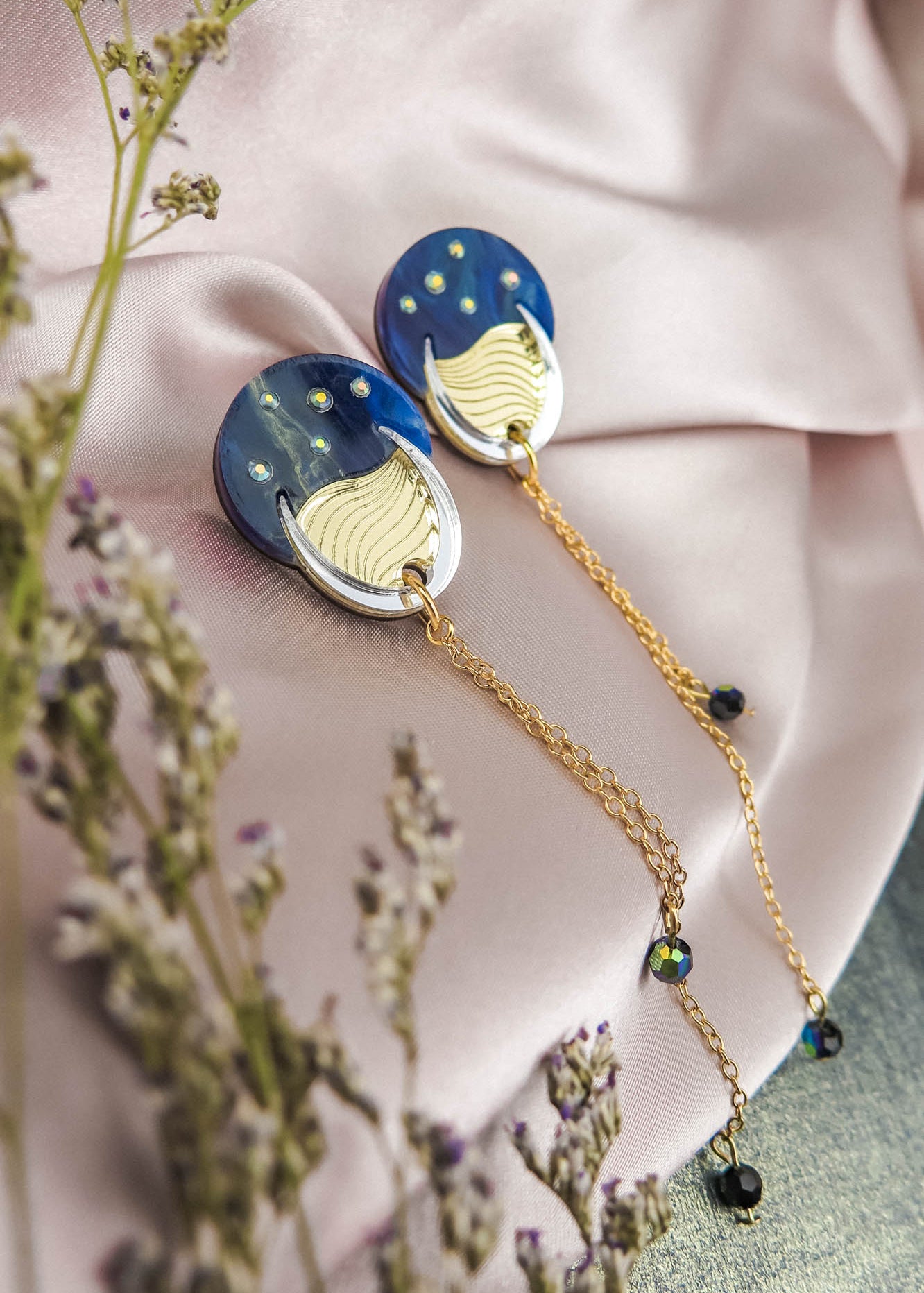 Two round, blue and gold earrings reminiscent of the night sky, with dangling gold chains, are placed on a light pink satin fabric. Small flowers with purple buds frame the foreground, adding to the festival magic of the scene.