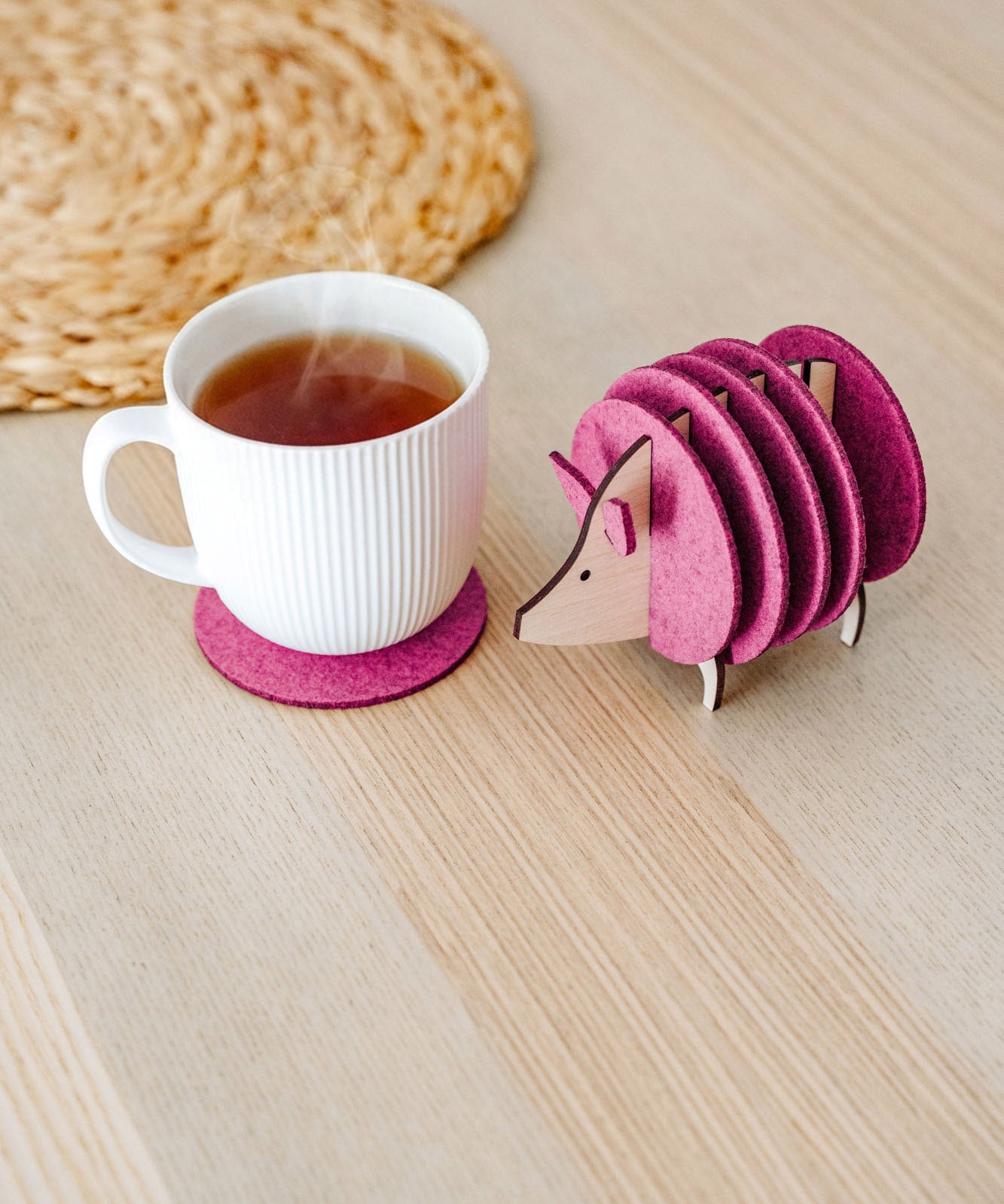 A white ribbed mug on a pink felt coaster sits next to a pink, hedgehog-shaped coaster holder on a light wooden table. A woven placemat is partially visible—perfect as a charming housewarming gift.