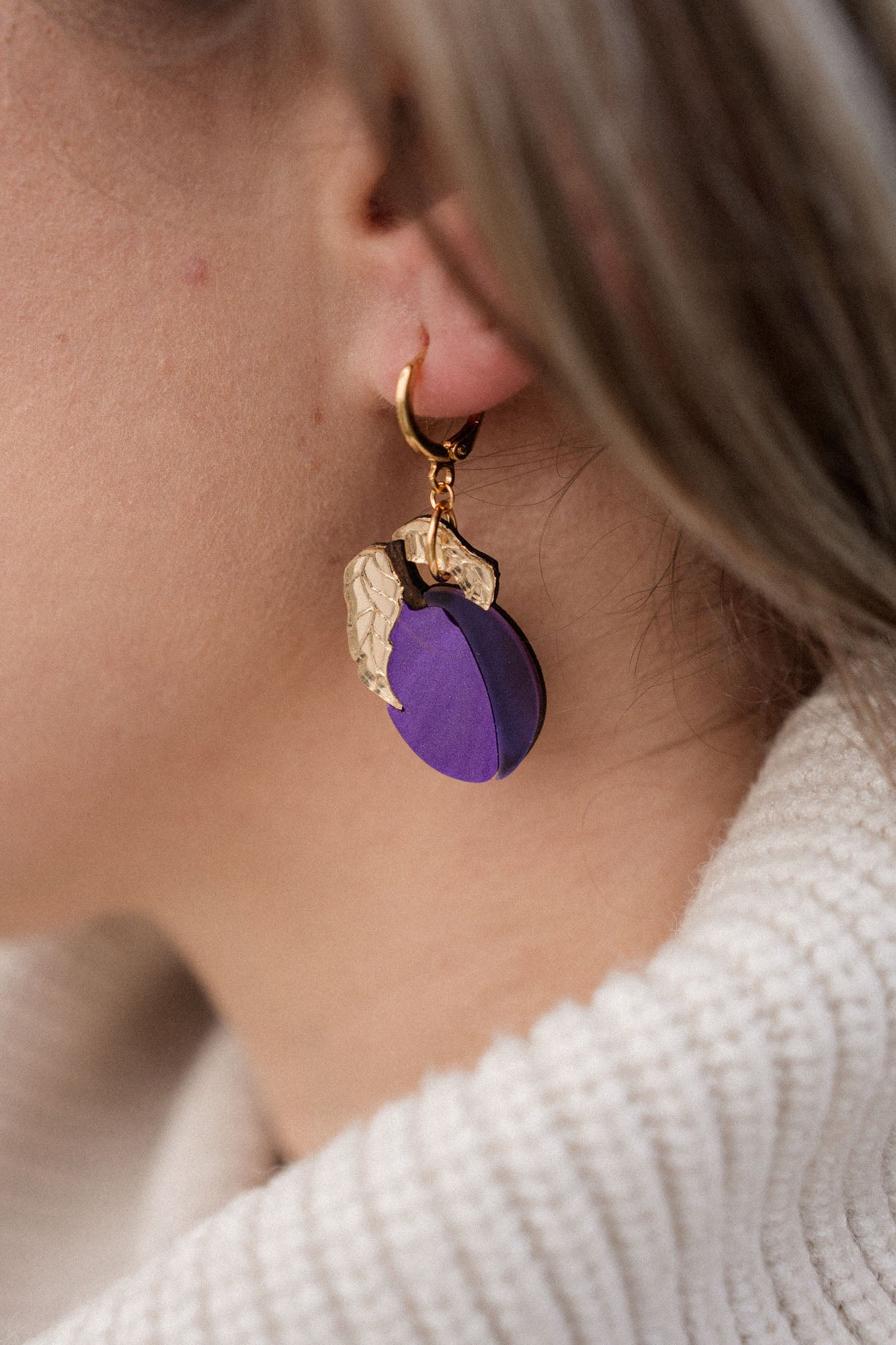 Close-up of a person wearing elegant purple plum earrings shaped like fruit with gold leaves. The person has light brown hair and is wearing a cream-colored, ribbed turtleneck sweater.