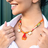 A woman smiling, wearing a white shirt and holding a handmade pomegranate flower necklace with gold and orange flower-shaped charms. She has small hoop earrings, rings on her fingers, and the necklace features a silk-print cord.