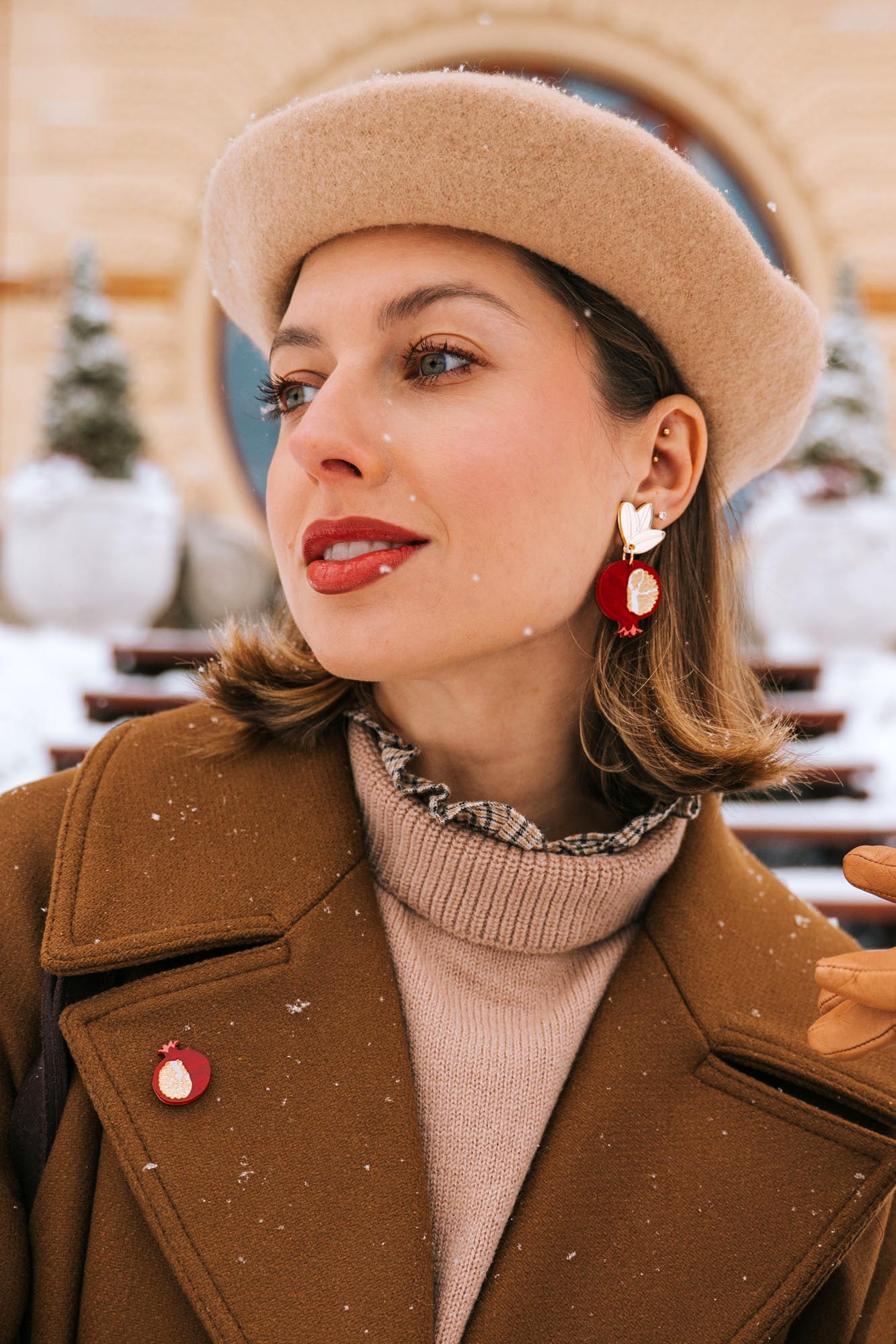 A woman in a tan beret and brown coat stands outside on a snowy day, wearing bold, handcrafted earrings with a bird design, a matching pin, tan gloves, and red lipstick. Snowflakes sprinkle her hair and coat.