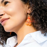 A woman with curly hair wearing a white shirt and handmade summer garden earrings smiles slightly while looking upward in natural light.