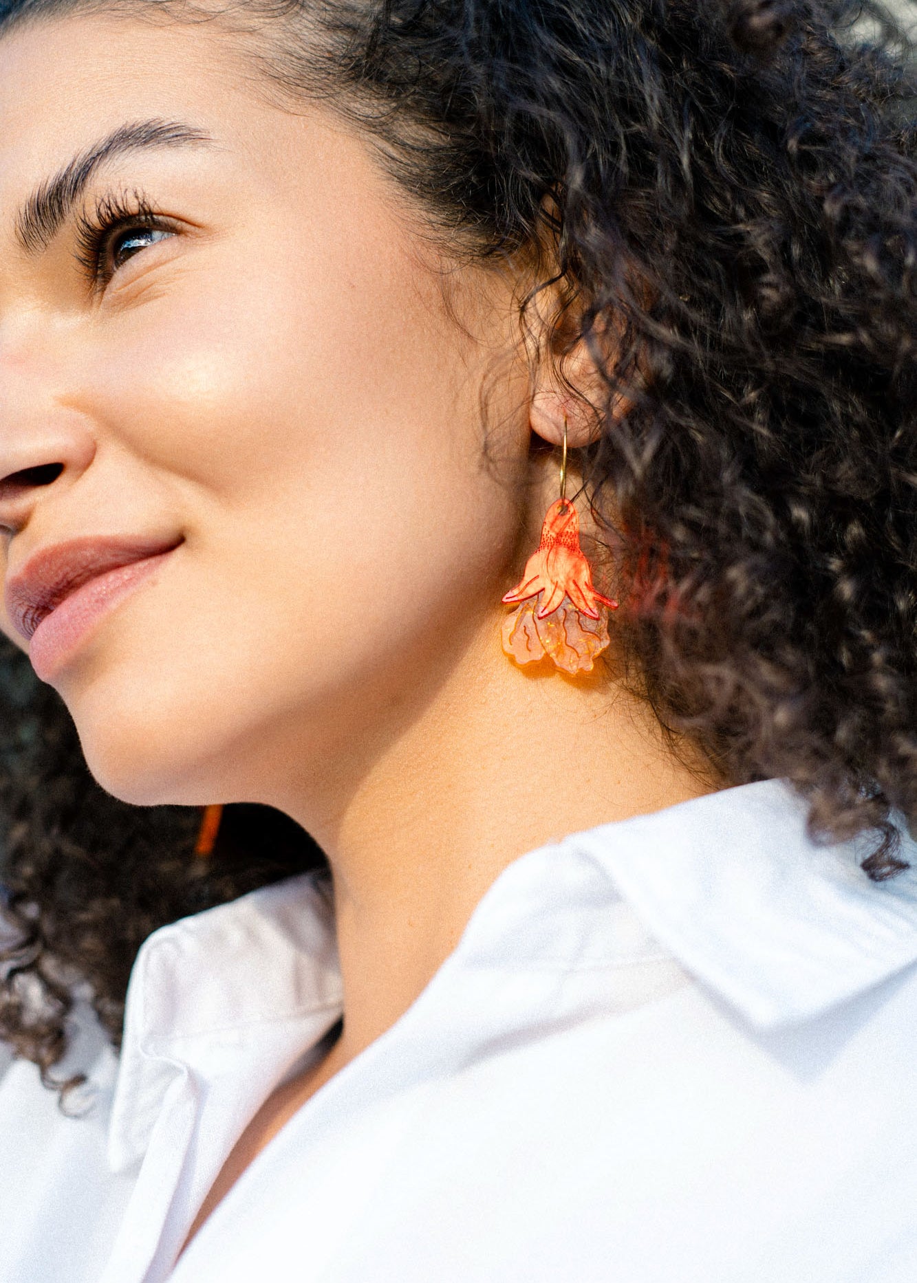 A woman with curly hair wearing a white shirt and handmade summer garden earrings smiles slightly while looking upward in natural light.