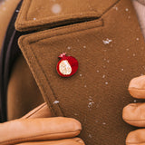 A person wearing a brown coat and tan gloves adjusts the lapel, which is decorated with a handcrafted Pomegranate Pin—a unique fashion accessory. Snowflakes are visible on the coat.