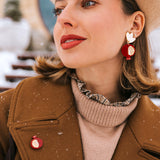 A stylish woman in a beige beret, brown coat, and tan gloves poses outdoors in snowy weather. She wears red and gold handcrafted earrings and a matching Pomegranate Pin fashion accessory, with snowflakes visible on her coat.