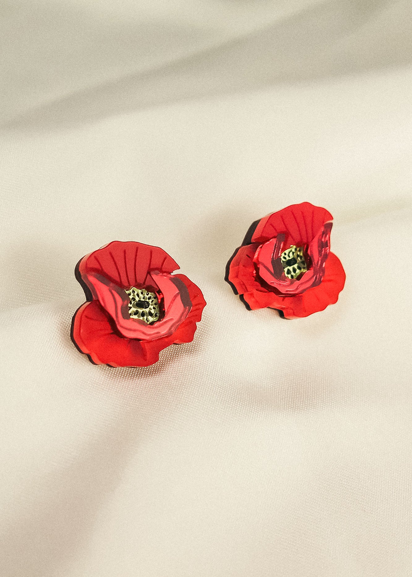 A pair of red, poppy design acrylic earrings with black and green centers, displayed on a soft, light-colored fabric background. Lightweight and eye-catching for any occasion.
